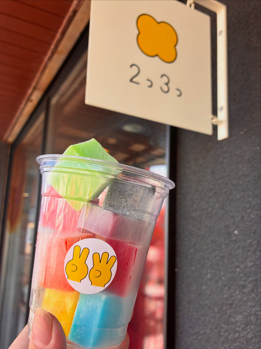 2つ3つの看板の下で持つ、カラフルな角切りの葛アイスが入ったカップ